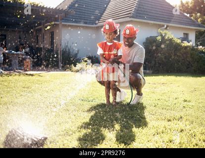 Father playing with his son outside. Little boy dressed as firemen. African American boy playing outside on a sunny day. Young male playing with a hosepipe in the garden. Dad and son spraying water f Stock Photo
