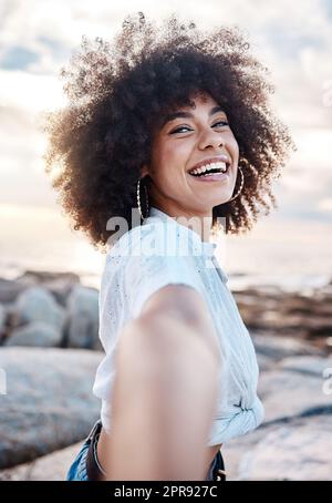 A young mixed race woman smiling on the beach while reaching out her hand. Happy hispanic female with a cool afro hairstyle outdoors Stock Photo