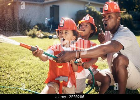 Little boys in firemen costumes. African American father playing with his children. Little boys playing in the garden. Boys spraying water from hosepipe outside. Excited brothers playing Stock Photo