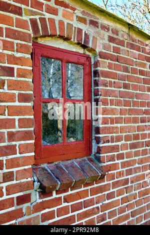 Old dirty window in a red brick home. Ancient casement with red wood frame in a historic building with lumpy paint texture. Exterior details of a windowsill in a traditional town or village Stock Photo