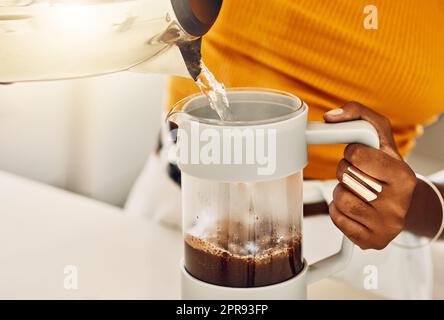 Female hands making morning coffee to enjoy with breakfast, pouring hot water from kettle in a kitchen. Closeup of casual, black woman preparing or brewing tea or espresso beverage at home. Stock Photo