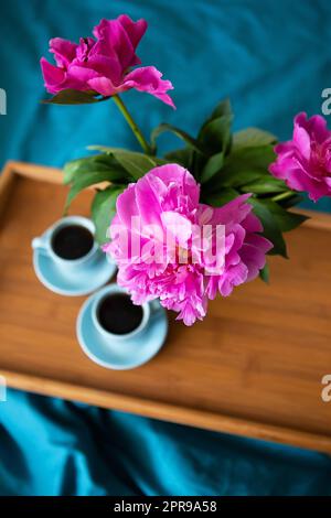 Beautiful pink peonies in a glass vase and two cups of coffee are standing on a wooden tray in the bed. Stock Photo