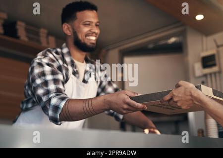 Heres your order, enjoy. a young man standing behind the counter in his restaurant and giving a customer her freshly made pizza. Stock Photo