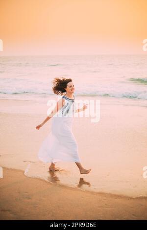 Goa, India. Young Caucasian Woman With Fluttering Hair In Wind In White Dress Walking Along Seashore, Enjoying Life And Smiling In Summer Sunlight Stock Photo