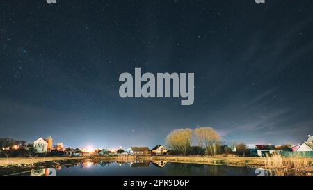 Belarus, Eastern Europe. Night Sky Stars Above Countryside Landscape With Lake Coast And Small Town Or Village. Natural Starry Sky Above Pond And Houses In Early Spring Night. Panorama Panoramic View. Stock Photo