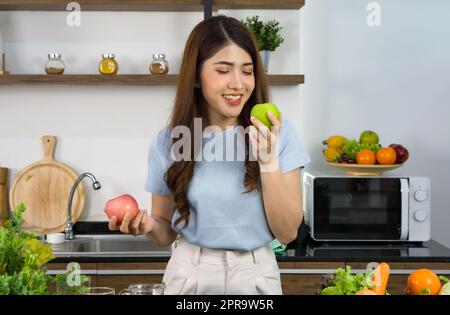 Young asian woman stand smiling,  hold red and green apple with both hands. Looking at the green one on the left. Stock Photo