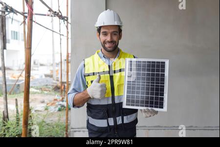 Young engineer in hardhat, safety vest and glove lift finger thumbs up while holding solar cell panel, present a source of energy to generate direct current electricity with a smile. Stock Photo