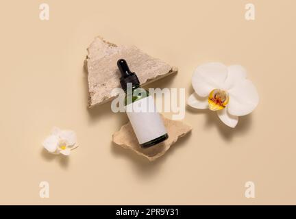 Green glass dropper bottle on stones near white orchid flowers on light yellow, Mockup Stock Photo