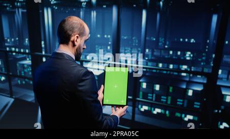 Data center technician holding tablet with green screen in server room, looking at isolated chroma key display. IT engineer inspecting mainframe computer processor, mockup template. Tripod shot. Stock Photo