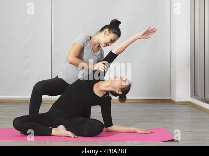 Instructor helping woman to do side bend at yoga class. Women sitting on mat in Easy seat pose or Sukhasana in the studio. Stock Photo