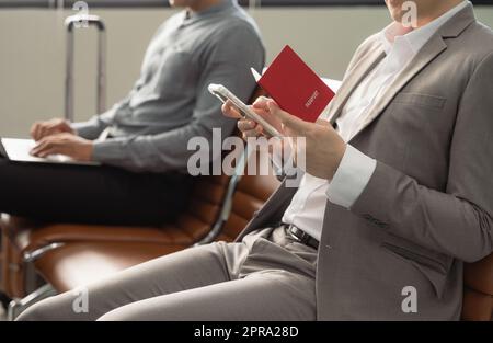 Businessman holding passport sitting at airport terminal and checking for his flight on mobile phone, waiting for plane and working while travelling. Stock Photo