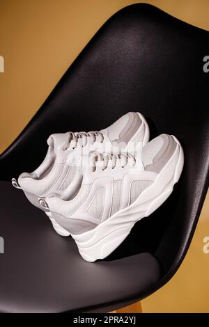 Close up of white sneakers leaning on back of black chair on beige studio background. Stock Photo