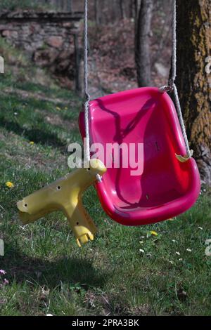 Broken swing on an abandoned playground with purple-yellow seat. The seat is broken. Grass and a tree are visible in the background. Stock Photo