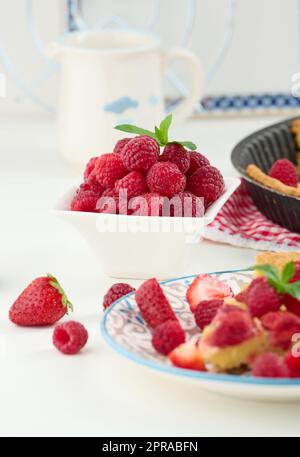 Ceramic white bowl with ripe raspberries and baked round cake on white table Stock Photo