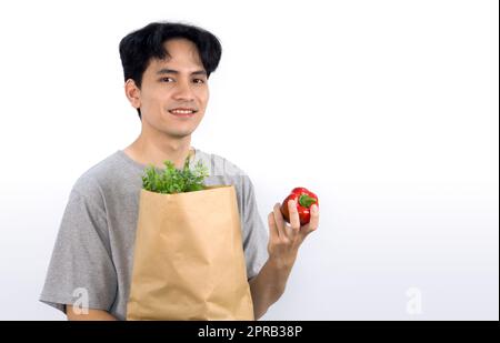 Young asian man in casual wear stand with a smile, holding the red bell pepper while carrying paper bag full with vegetable. Portrait on white background with studio light. Stock Photo