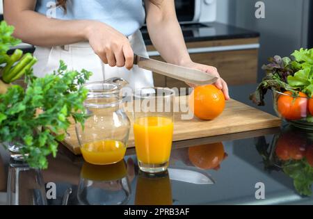 Closeup hand holding knife cutting orange fruit on a wooden chop board. Jar with mixed fruit juice  and glass bowl with a variety of fruits and vegetables is placed on the kitchen counter. Stock Photo