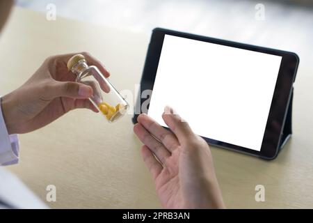 Back view of pharmacist in white gown sit at desk holding glass pill packer bottle with wooden cap in front of blank screen tablet computer. Stock Photo