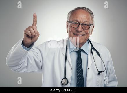 Ah yes Ive got a great idea. an elderly male doctor holding his finger up in a studio against a grey background. Stock Photo