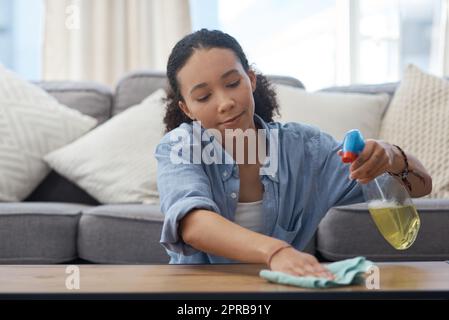 Caring for my expensive table is a joy. a young woman wiping down her wooden coffee table in her living room at home. Stock Photo