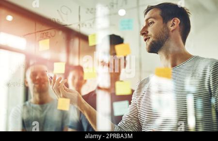 Group of young designers brainstorm as a team. Writing down new ideas on glass board inside workplace together. Business people meeting at the office to discuss notes and share creative designs. Stock Photo