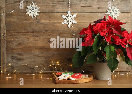 Poinsettia (traditional Christmas flower), cookies and string lights on wooden table. Space for text Stock Photo
