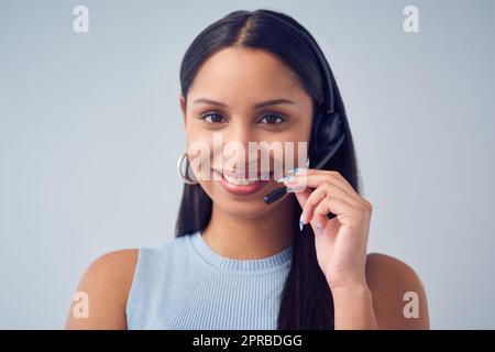 How can I hep you today. Cropped portrait of an attractive young female call center agent standing against a grey background in studio. Stock Photo