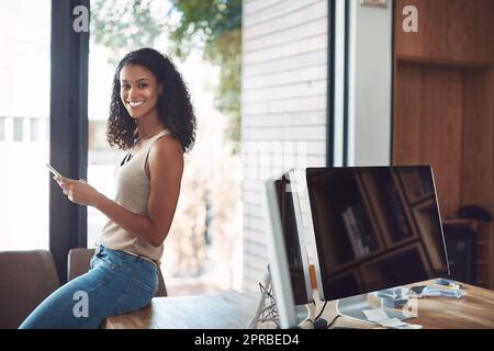 Smiling, modern and casual female in her home office happy to be working remote. Portrait of a confident young online worker sitting on a desk. Digital marketing employee with a smile holding a phone