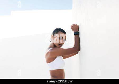 Portrait of a fit female leaning white wall and looking straight at a camera. Fitness influencer taking a short break during a workout. Stock Photo