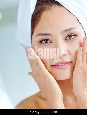 Freshly-showered radiance. A beautiful young Asian woman touching cheeks while wearing a towel on her hand. Stock Photo