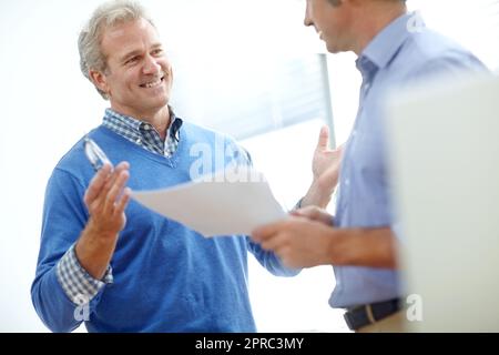 Project details they can both agree on. two positive-looking mature businessmen standing in an office and discussing paperwork. Stock Photo