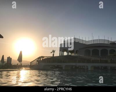 A large stone building near the water, a pool against the blue sky and a big sun, a sunset in the evening in a tropical seaside resort. Stock Photo