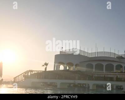 A large stone building near the water, a pool against the blue sky and a big sun, a sunset in the evening in a tropical seaside resort. Stock Photo