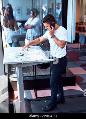 Burning some calories while making some deals. a young businessman talking on a cellphone and going through paperwork while walking on a treadmill in an office. Stock Photo