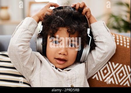 Im having a jam packed day. Portrait of an adorable little boy listening to music on headphones while sitting on a sofa at home. Stock Photo