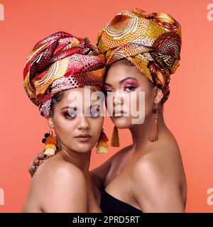 Wear your history and heritage with pride. Studio shot of two attractive young women wearing traditional African head wraps posing together against an orange background. Stock Photo