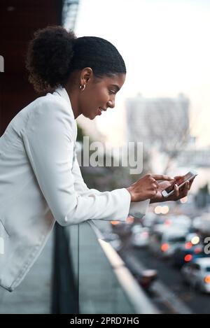 Relaxed business woman texting on her phone while standing on a balcony on her lunch break. Young corporate female browsing the internet or scrolling on social media after work in a city Stock Photo