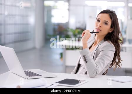 Confident businesswoman thinking about a corporate strategy to boost sales at her desk while holding a pen with her laptop open in an office. Successful professional female planning a campaign alone Stock Photo