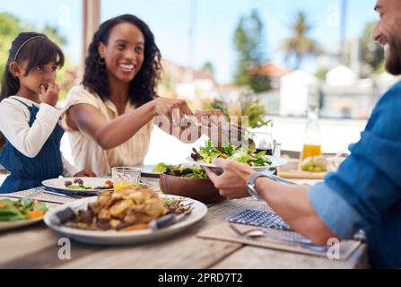 She always makes sure we maintain a healthy diet. a beautiful young woman dishing up salad on her husbands plate while enjoying a meal with family outdoors. Stock Photo