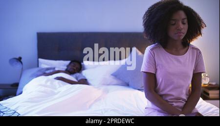 I have a big decision to make. a concerned looking young woman sitting on a bed with her husband sleeping in the background. Stock Photo