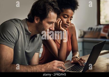 Diverse couple searching for a new rental apartment home on their laptop sitting together in their kitchen at their house. Man and woman in an interracial marriage relationship doing online shopping Stock Photo