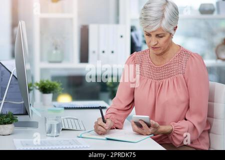 Shes having a busy day at the office today. a mature woman writing down details from her cellphone to her diary in her office at work. Stock Photo
