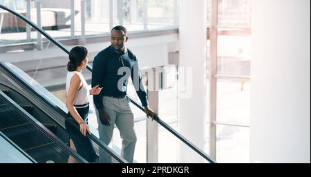 Keeping the discussion moving. two young businesspeople having a chat while going down an escalator in a modern workplace. Stock Photo