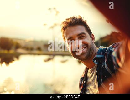 Its awesome out here. Cropped portrait of a handsome young man taking selfies while spending the day at a lake. Stock Photo