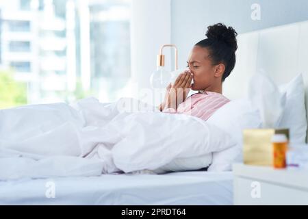 Guess Ill be stuck in bed today. a sickly young woman blowing her nose with a tissue in bed at home. Stock Photo