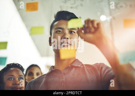 A marketing professional brainstorming ideas with colleagues, writing on transparent board with sticky notes during meeting. Young, Asian entrepreneur discussing a work project schedule or timeline. Stock Photo