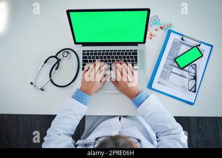 Technology greatly influences medical processes and practices. High angle shot of an unrecognizable doctor using a laptop and cellphone with a green screen in his office. Stock Photo
