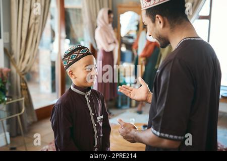 Muslim father and son talking and bonding in the living room during a religious holiday at home. Cute, smiling and happy Islamic boy listening to advice from his dad while wearing traditional outfit Stock Photo