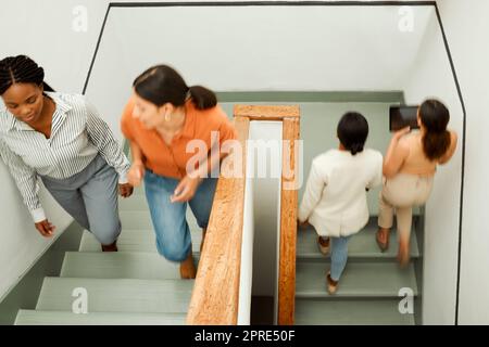 Overhead of busy, diverse female colleagues returning to work after break, with blurred digital tablet screen. Active business woman in motion talking and walking up office building stairs. Stock Photo