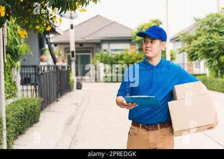 Asian young delivery man courier in uniform hold parcel post boxes service shipment and walking finding and looking for customer home location, Online Stock Photo