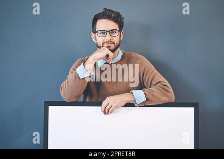 Young man standing behind an empty sign with copyspace. Smiling, attractive and smart male holding a blank board announcement for marketing. Handsome guy with a poster for an advert, banner or news. Stock Photo